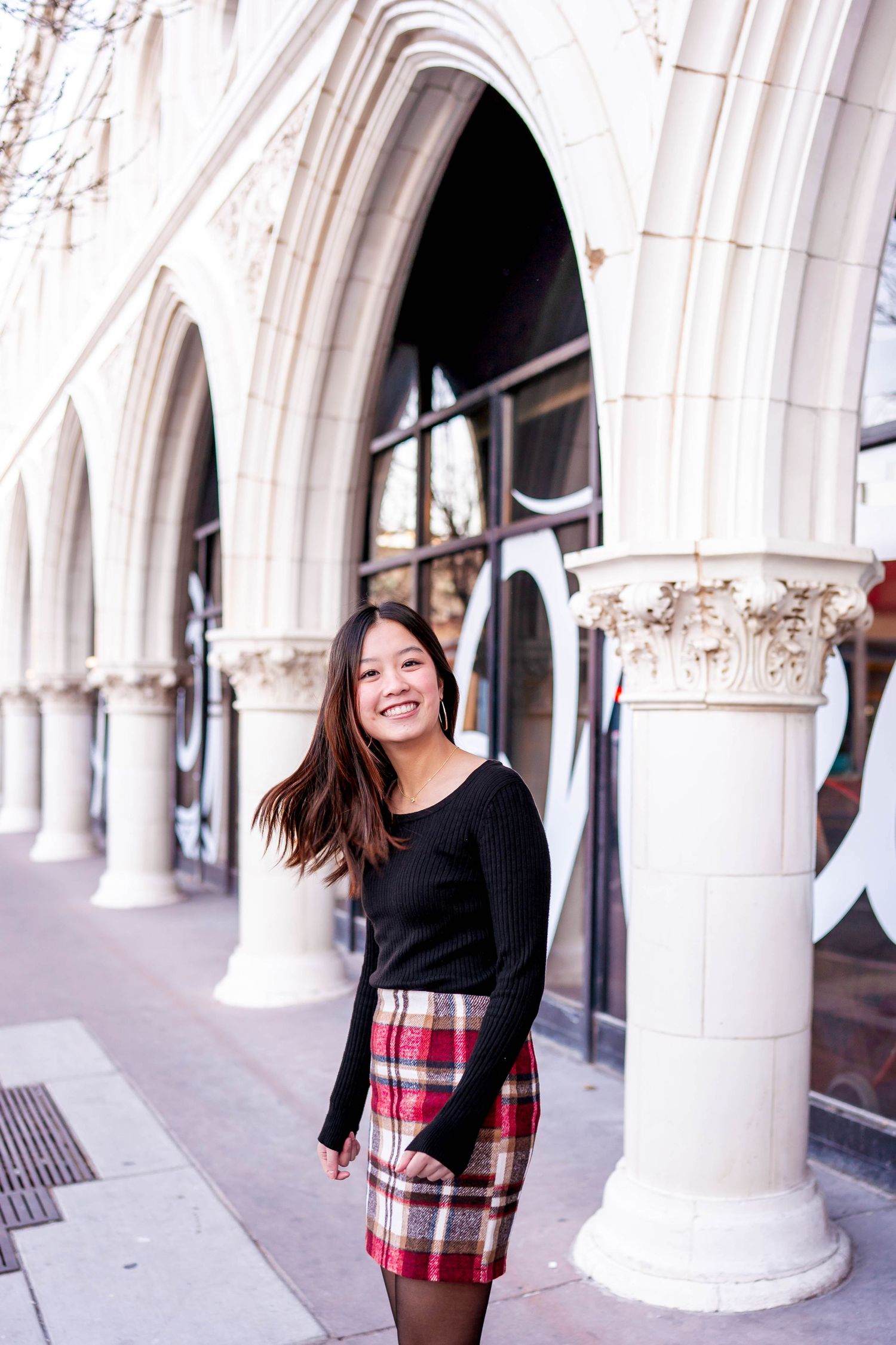 Person in plaid skirt and black sweater poses by ornate white stone archways.