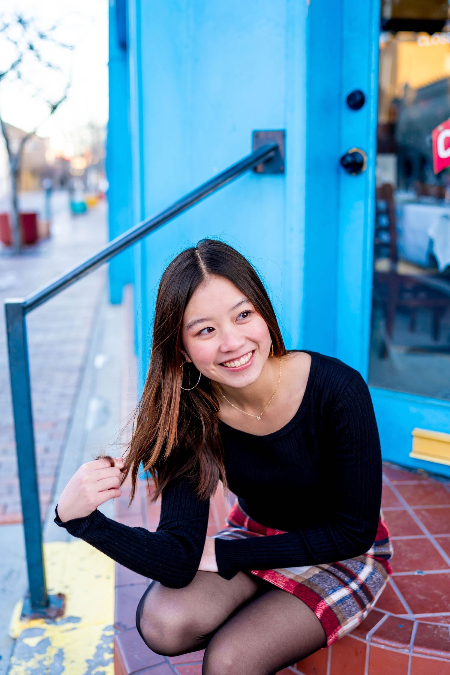 Person in black sweater and plaid skirt sits by bright blue storefront.