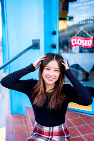 Person in black sweater and plaid skirt poses playfully near blue building.