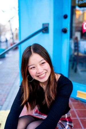 Person in dark sweater and plaid skirt smiles while sitting near blue storefront.
