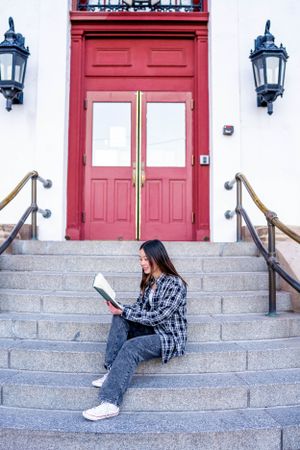 Person reads book while sitting on stone steps with red door and lanterns in background.
