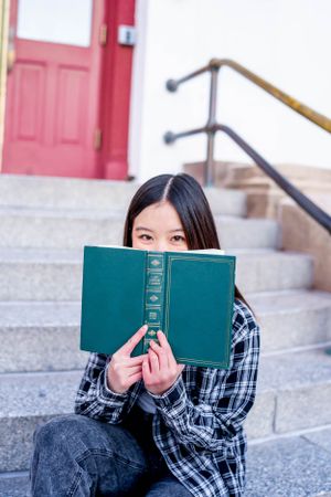 Student sits on stone steps reading green book by red door entrance.
