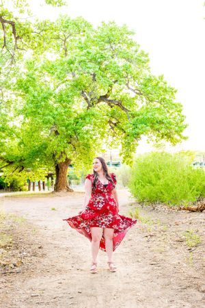 Red floral dress twirling on dirt path under green tree canopy.