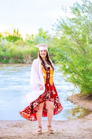 Graduate in red dress and cap posing by riverside with green foliage.