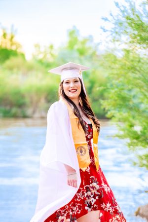 Graduate in red floral dress and white graduation gown poses outdoors by water.