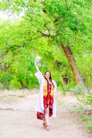 Graduate celebrates in white cap and gown with red floral dress on nature path.