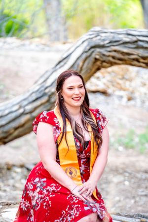 Graduate in red floral dress and gold sash sits on fallen log in natural setting.