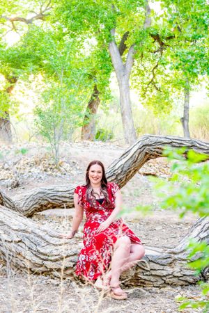 Graduate in red floral dress poses on curved fallen tree trunk surrounded by greenery.