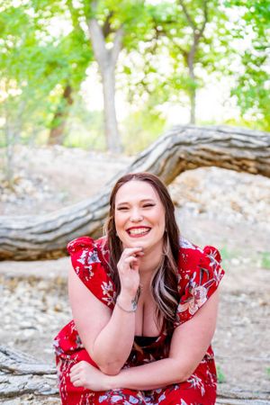 Graduate in red floral dress smiles while sitting outdoors with natural backdrop.