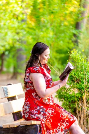 Graduate in red floral dress reads book while sitting on park bench.