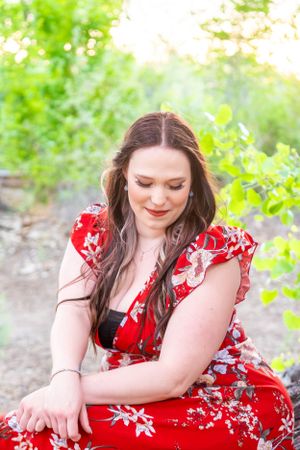 Graduate in red floral dress poses thoughtfully in outdoor garden setting.