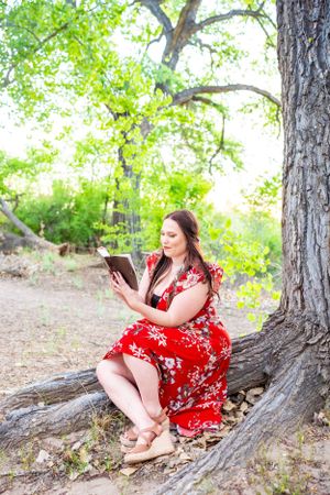 Graduate in red dress reads while sitting against tree trunk in natural setting.