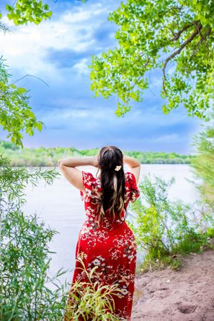 A woman in a flowing red floral dress stands by a lake surrounded by green trees on a sunny day.