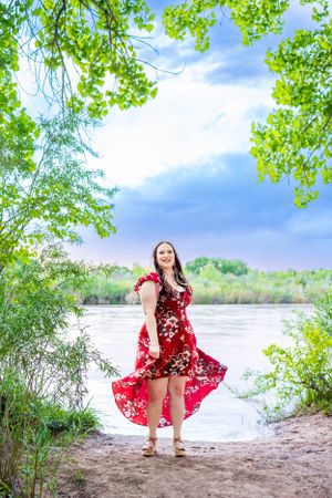 A red floral dress flows in the breeze as someone poses by a serene lakeside under green trees.