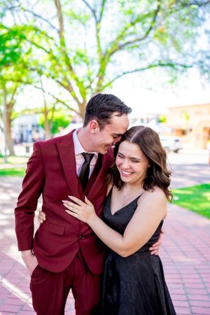 A couple shares an intimate moment outdoors, with one wearing a burgundy suit and the other in a black dress.