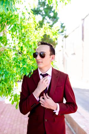 Person in a burgundy suit and sunglasses poses confidently against a leafy green background.
