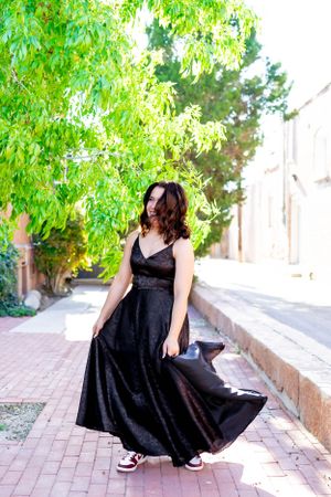 Person in a black formal dress poses on a brick pathway under green trees.