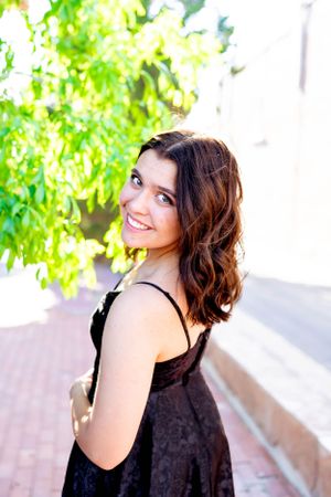 A person in a black dress smiles over their shoulder on a sunny brick pathway.
