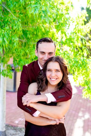 A couple embraces outdoors, wearing coordinating burgundy and black formal attire.