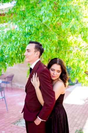 A romantic portrait of a couple in burgundy and black formal attire against lush greenery.