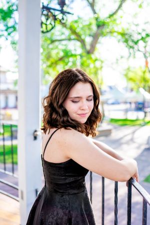 A woman in a black dress poses elegantly on a porch with sunlit trees in the background.