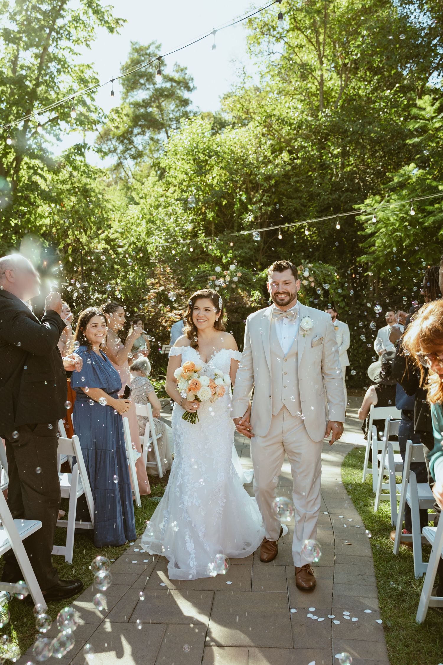 Wedding party processes down an outdoor garden path decorated with string lights and greenery.