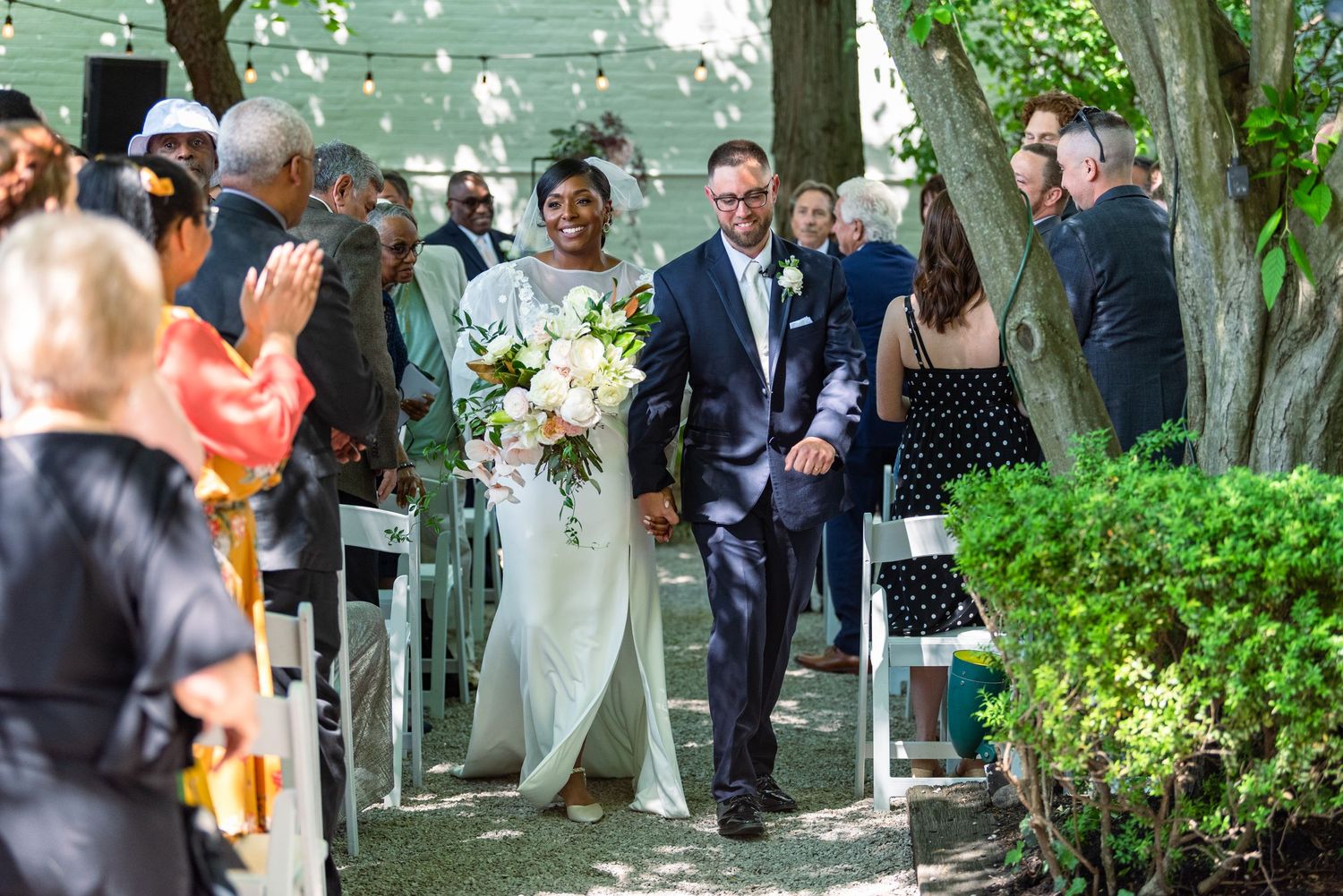 Wedding guests gather outdoors under trees during a garden ceremony celebration.