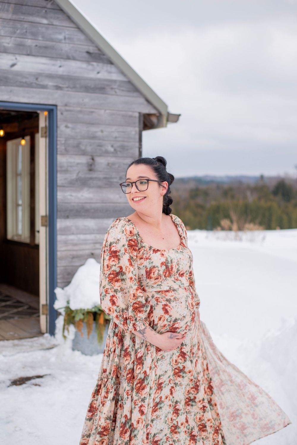 A person in a floral dress and glasses stands outside a rustic wooden cabin in the snow.