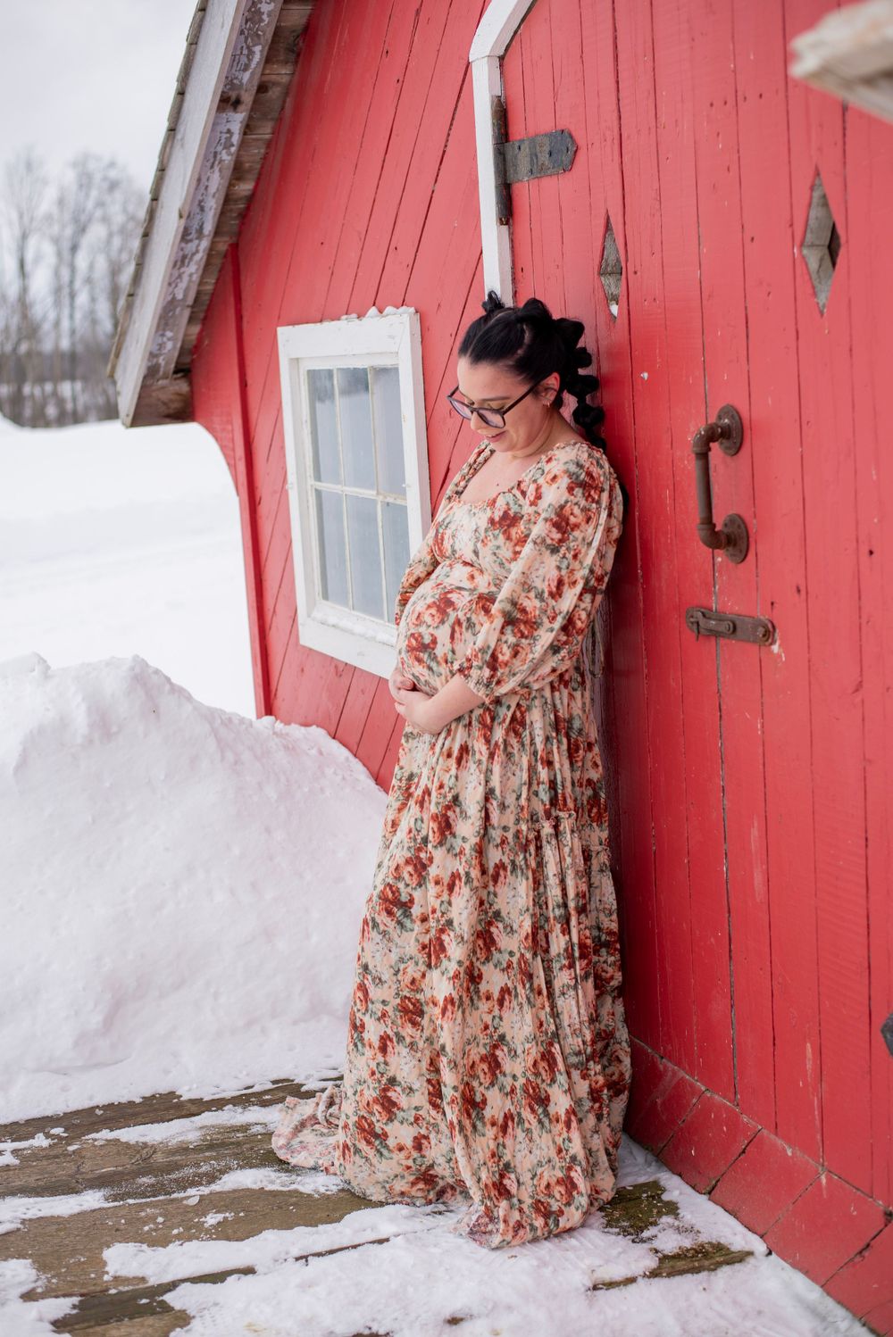 Winter maternity portrait against a bright red barn wall with snow on the ground features a floral maxi dress.