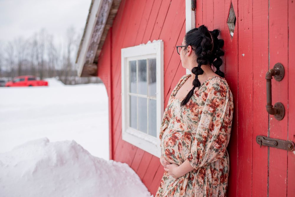 A pregnant woman in a floral dress stands beside a red barn door in a snowy winter landscape.