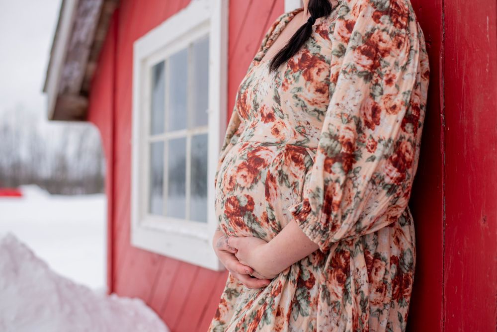 A pregnant person in a floral dress stands against a red barn wall during winter, with snow visible in the background.