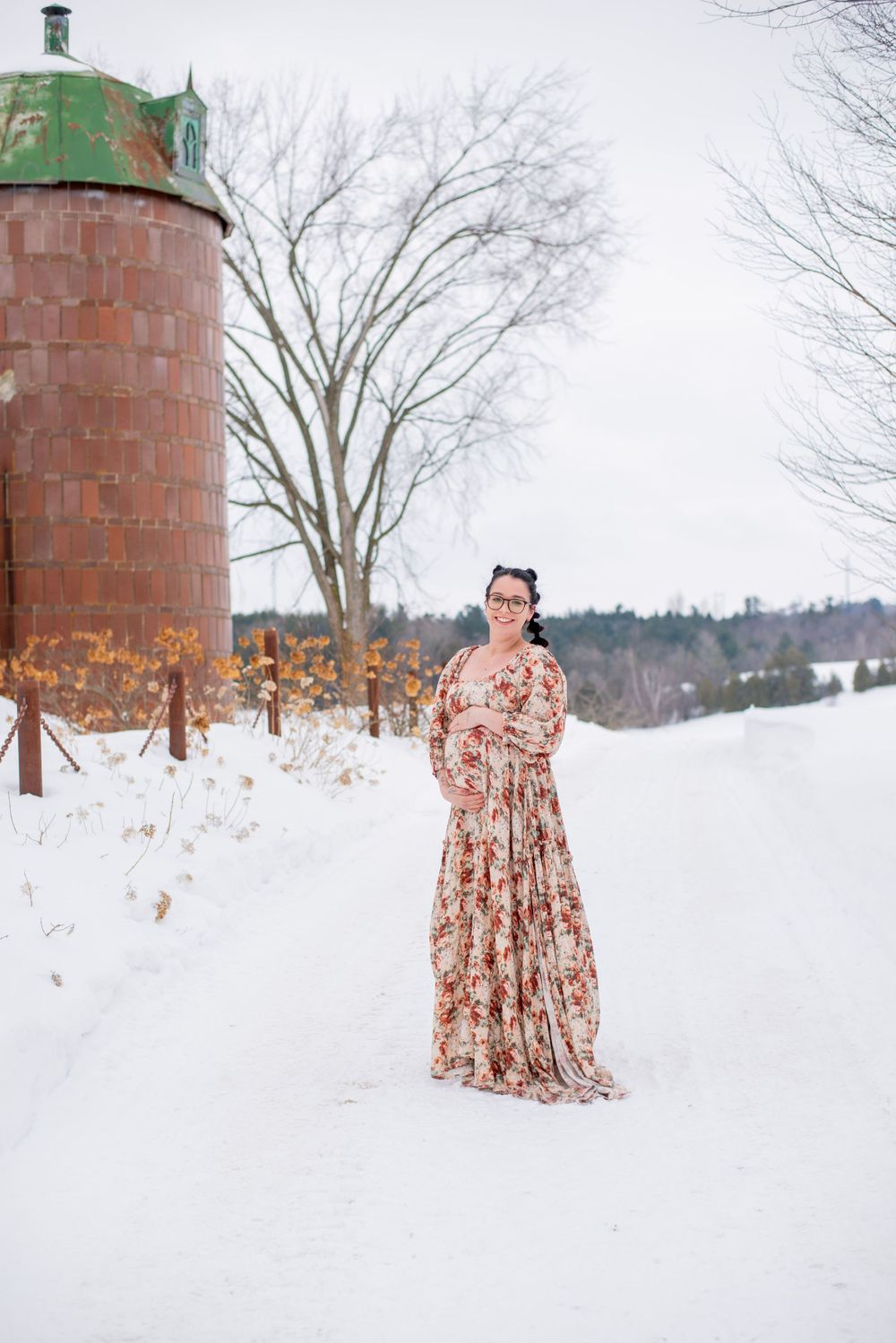Person in floral maxi dress poses in snowy landscape with rustic red silo during winter photoshoot.