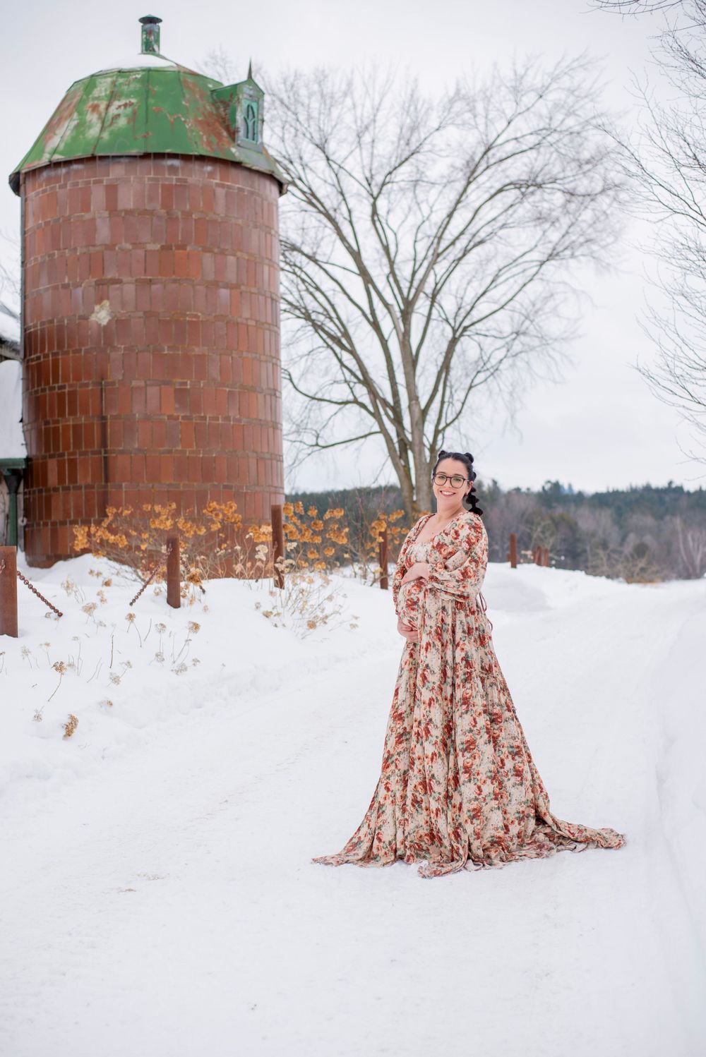 A person in a floral maxi dress poses in front of a rustic farm silo amidst a snowy winter landscape.