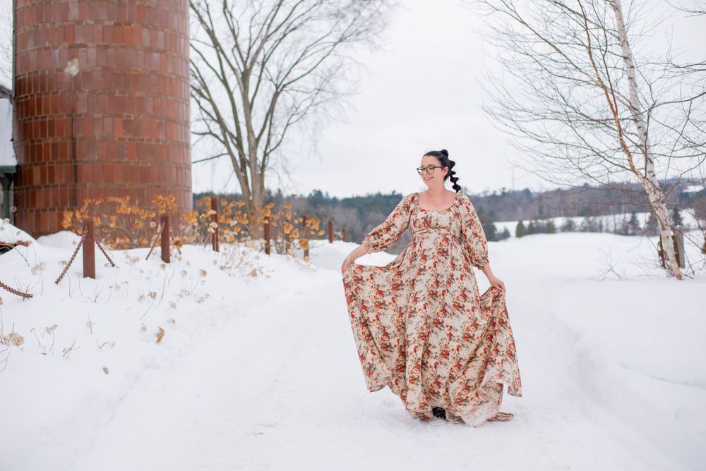 A floral dress billows in the winter breeze against a snowy rural landscape with a rustic brick silo in the background.