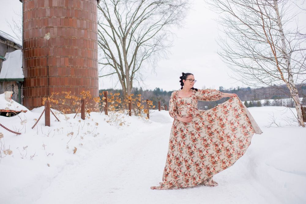A flowing floral dress swirls dramatically against a snowy winter landscape with a rustic brick silo in the background.