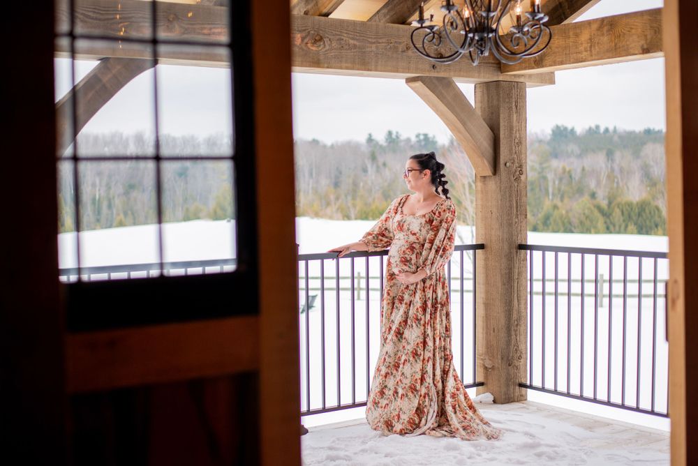 A person in a floral gown stands on a snow-covered balcony with mountain views while holding the railing.