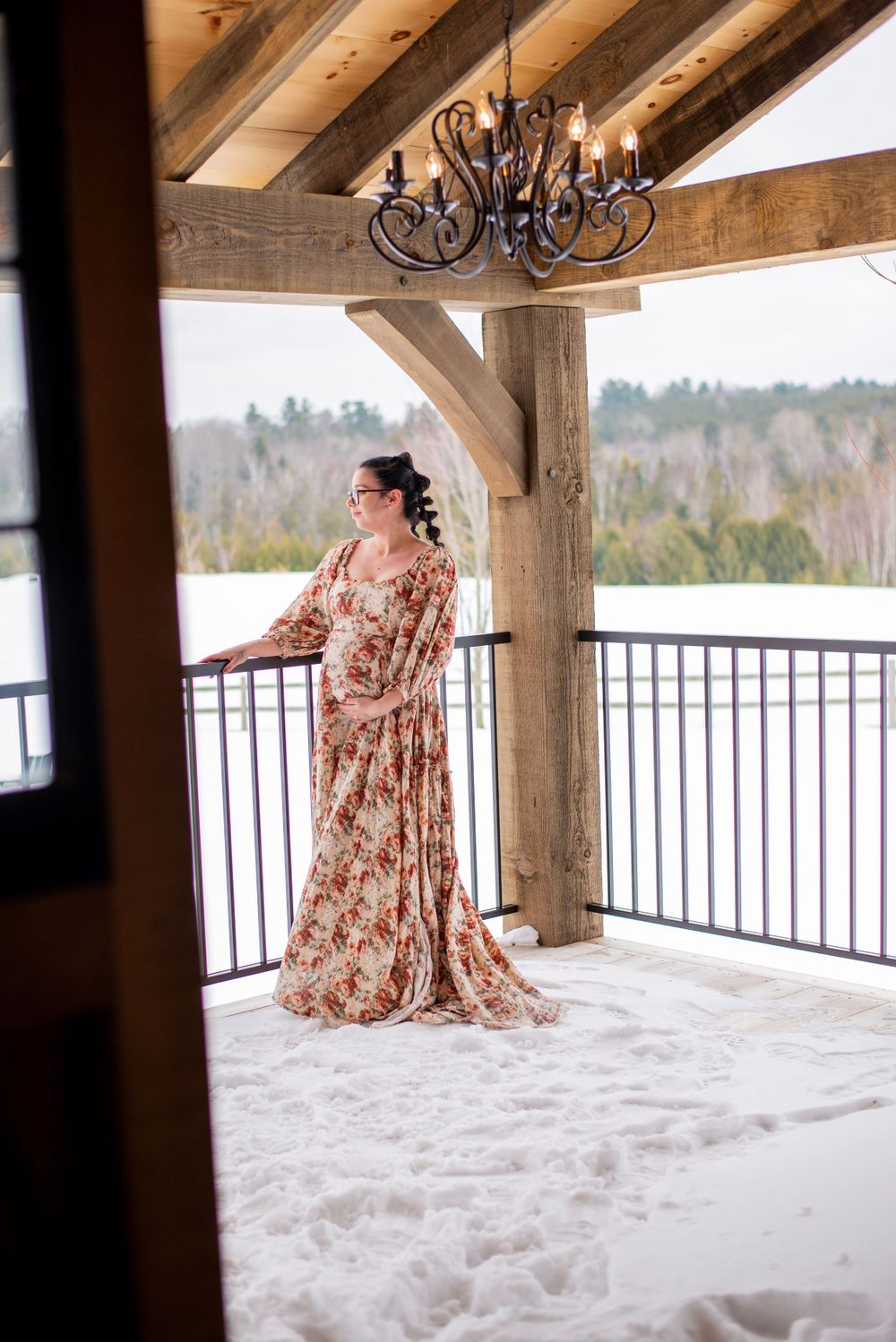 A rustic covered porch with wooden beams and an ornate chandelier overlooking a snowy winter landscape.