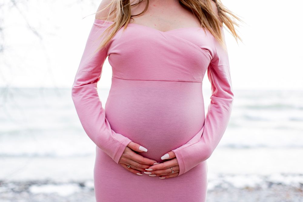 Person wearing a fitted pink long-sleeve maternity dress cradles their pregnant belly against a bright background.