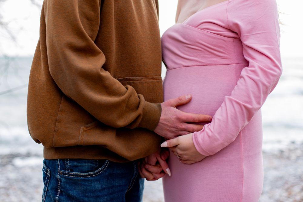 Cropped view of couple holding hands, with person in pink maternity dress and brown sweater visible.