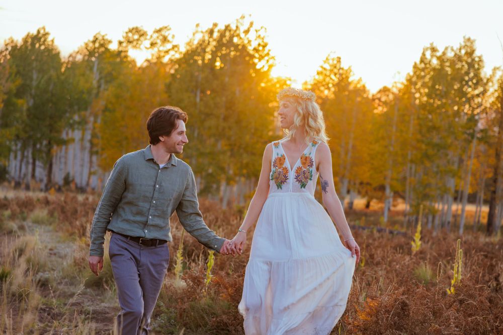 Couple holding hands walking through golden autumn forest at sunset in a floral white dress and green linen dress shirt.