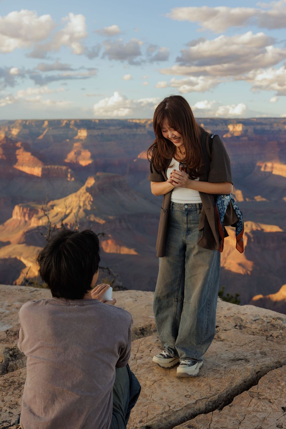 Couple at Grand Canyon overlook, woman smiling while man proposes to her at sunset.