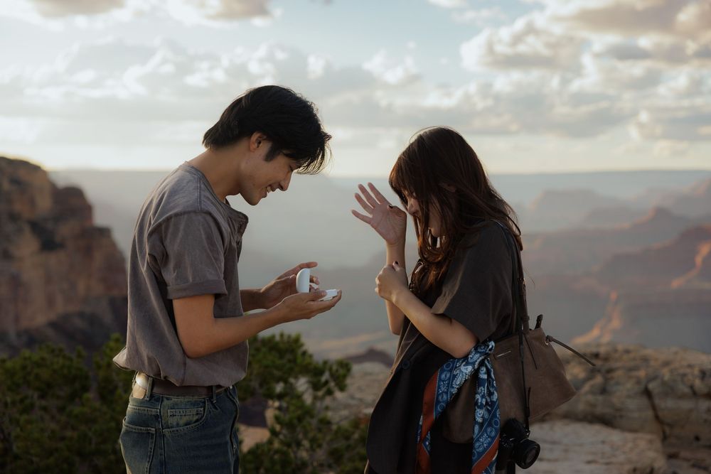 A couple shares a romantic moment at the Grand Canyon rim during sunset, as a man presents a ring box to a surprised woman.