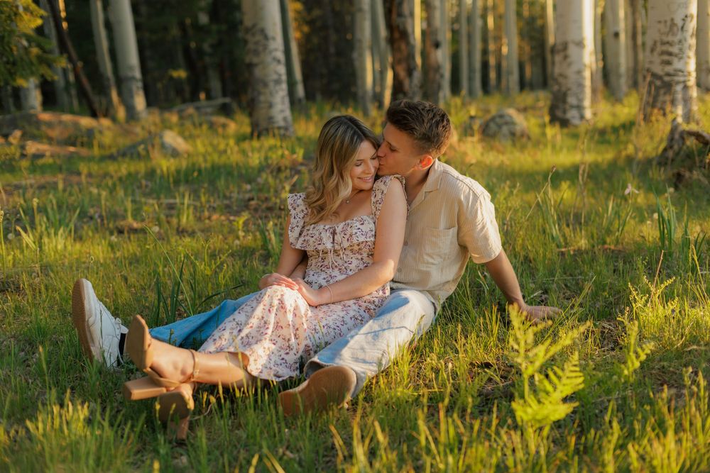 Couple sitting together in a sunlit forest meadow, woman in floral dress and man in cream shirt sharing a romantic moment.