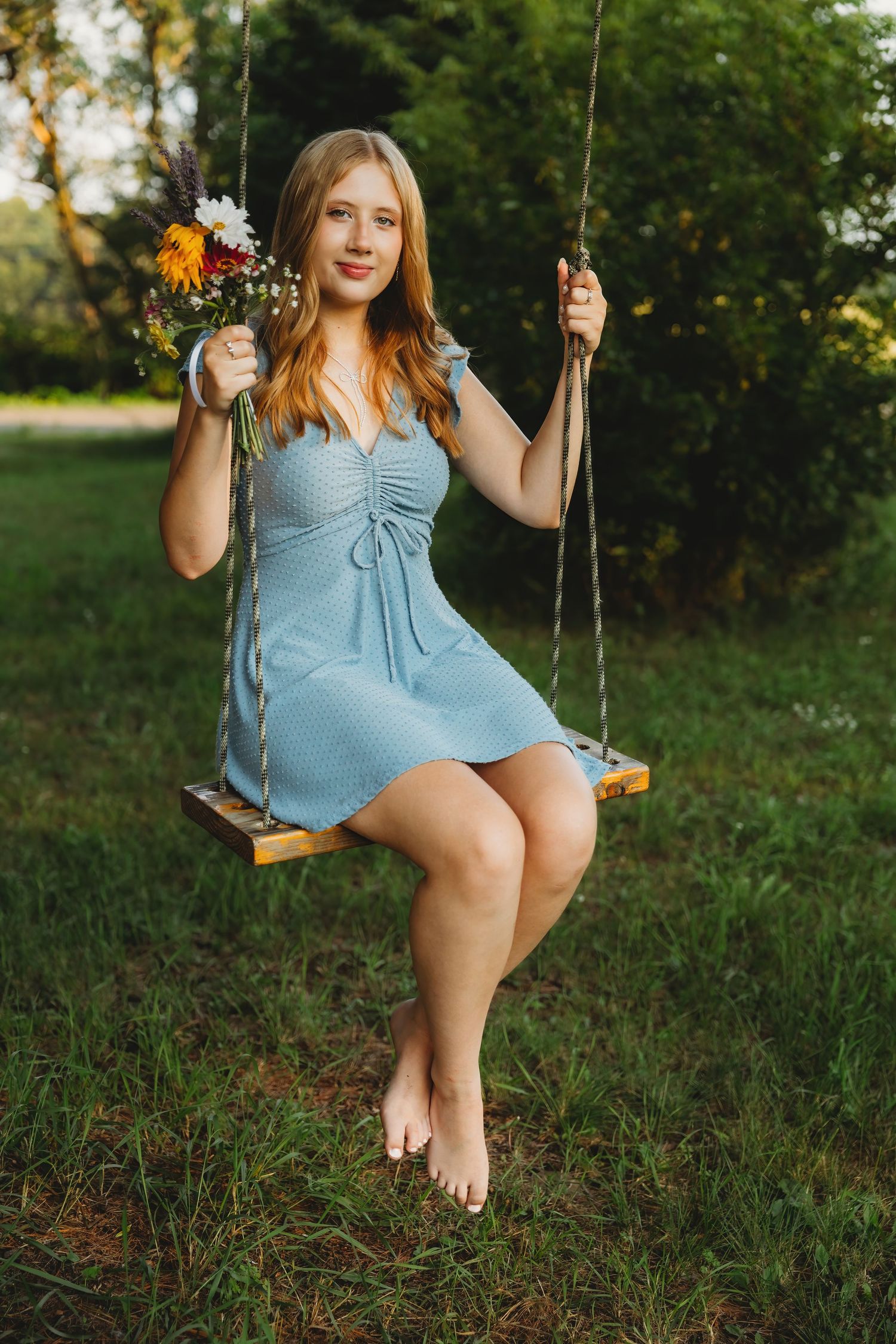 Woman in blue dress sitting on wooden swing holding flowers in garden setting.