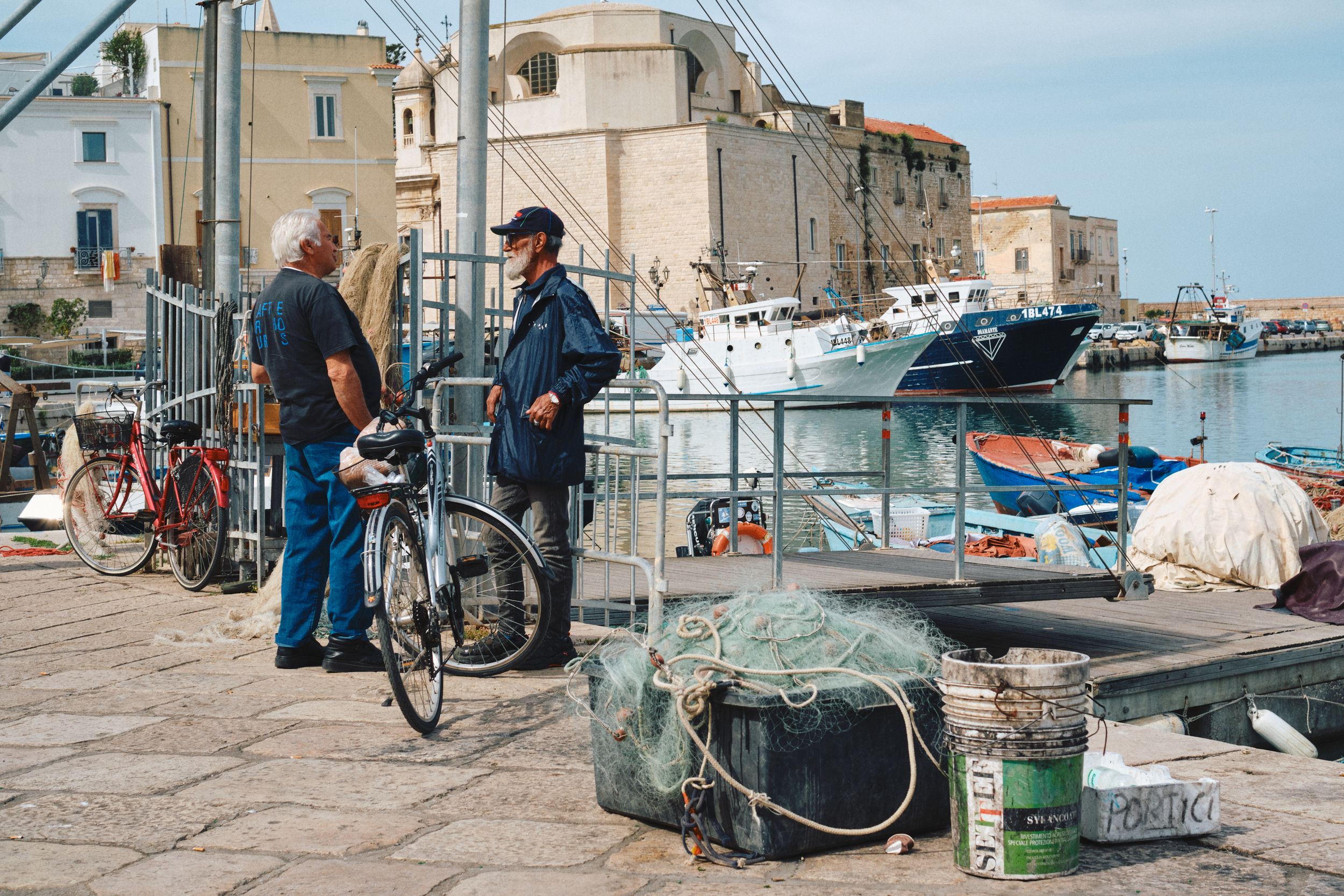 People conversing on a waterfront dock with fishing boats in the background.