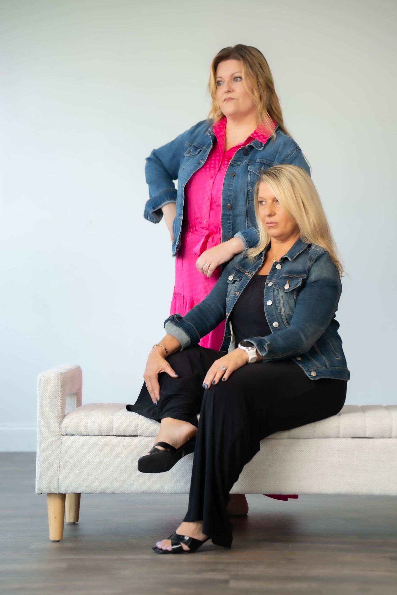 Two women relax on a white couch wearing casual denim and black outfits.