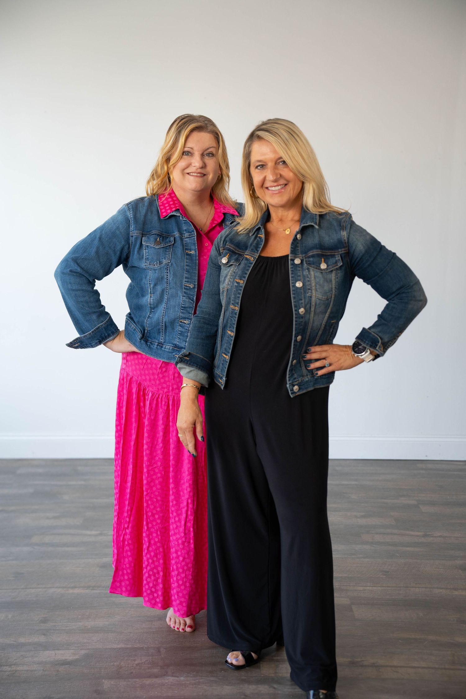 Two women pose in coordinating outfits featuring denim jackets and contrasting pink and black dresses.