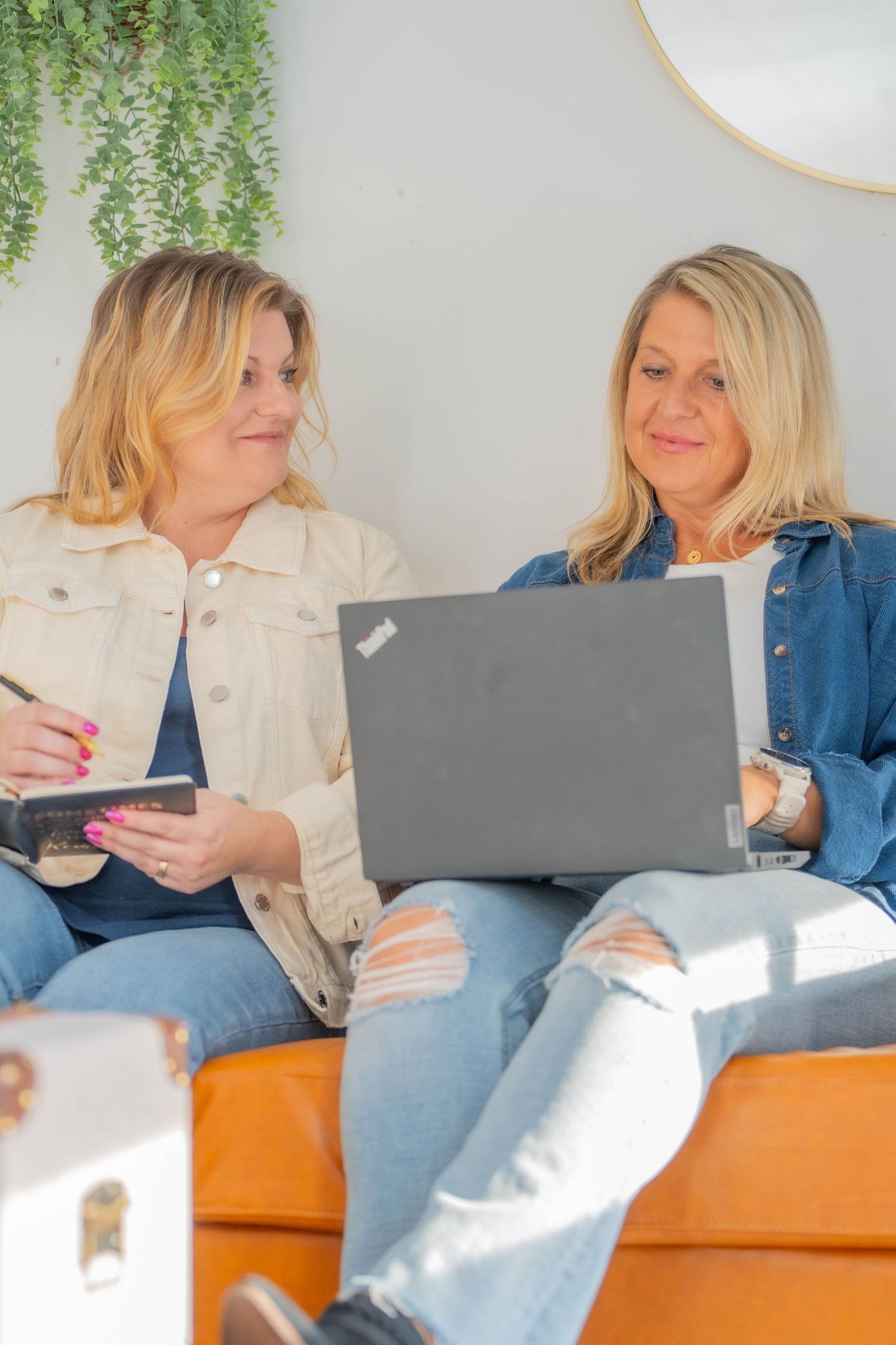 Two women sitting on orange couch working together on laptops in sunny room.