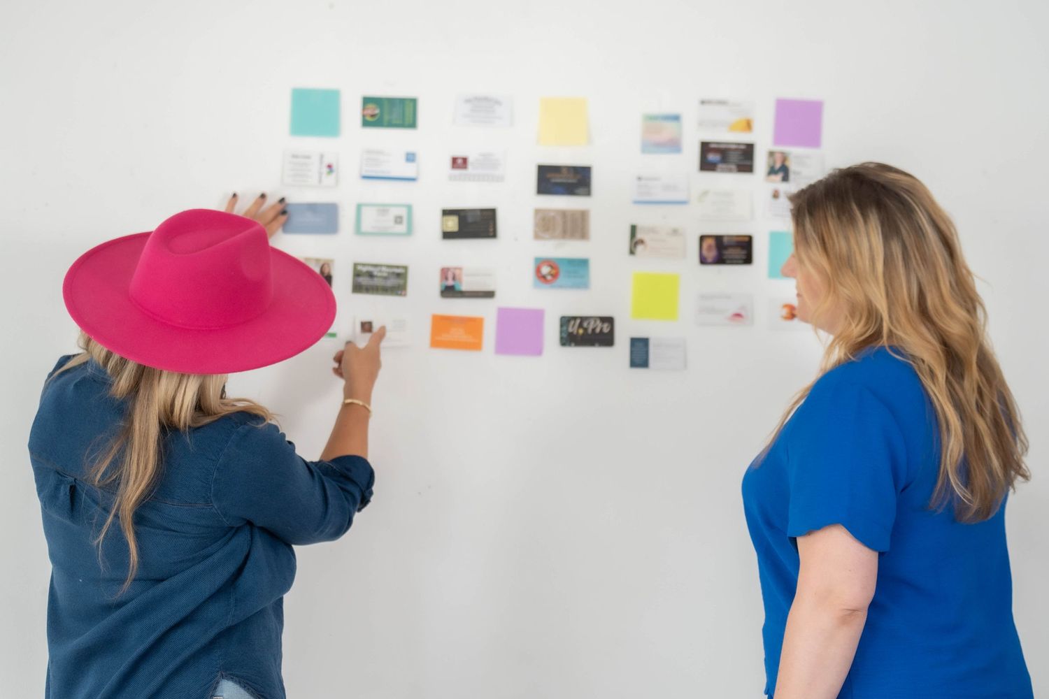 Woman in denim jacket reviewing colorful sticky notes and papers on white wall.