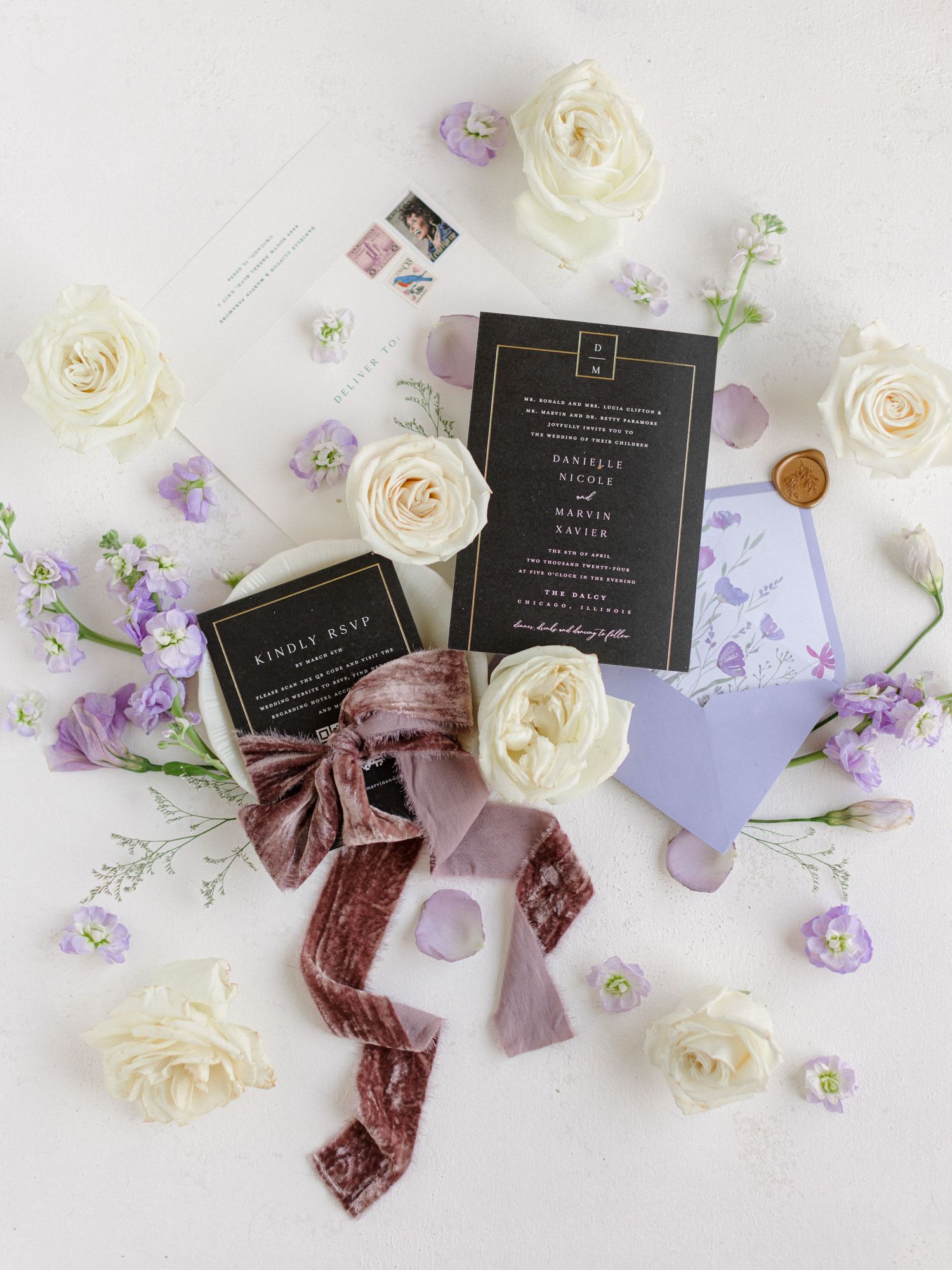 Wedding stationery flatlay with white roses, purple flowers, and velvet ribbon on light background.
